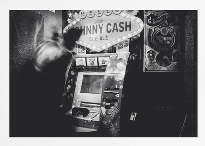 A grainy, black and white photograph of a person blurred by motion standing in front of a Johnny Cash themed slot machine. A lit marquee sign above the machine reads "HELLO I'm JOHNNY CASH OLÉ OLÉ". The setting appears to be a dark bar or casino. Wall Art