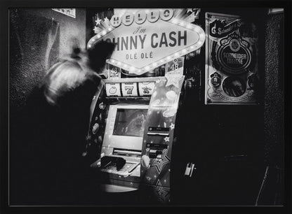 A grainy, black and white photograph of a person blurred by motion standing in front of a Johnny Cash themed slot machine. A lit marquee sign above the machine reads "HELLO I'm JOHNNY CASH OLÉ OLÉ". The setting appears to be a dark bar or casino. Wall Art