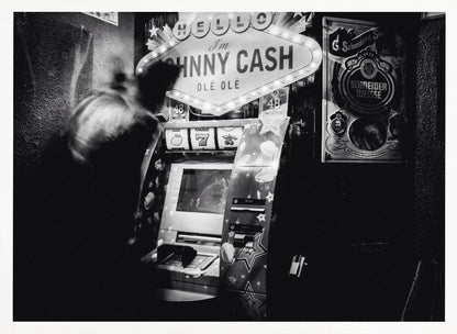 A grainy, black and white photograph of a person blurred by motion standing in front of a Johnny Cash themed slot machine. A lit marquee sign above the machine reads "HELLO I'm JOHNNY CASH OLÉ OLÉ". The setting appears to be a dark bar or casino. Wall Art