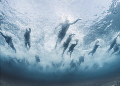 An underwater photograph looking up at a group of swimmers competing in a race. Their bodies are silhouetted against the bright surface of the turbulent blue water, captured mid-motion amidst a flurry of bubbles and foam. Print