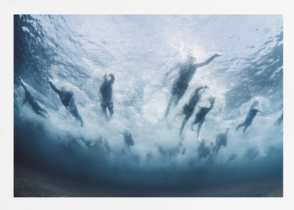 An underwater photograph looking up at a group of swimmers competing in a race. Their bodies are silhouetted against the bright surface of the turbulent blue water, captured mid-motion amidst a flurry of bubbles and foam. Print