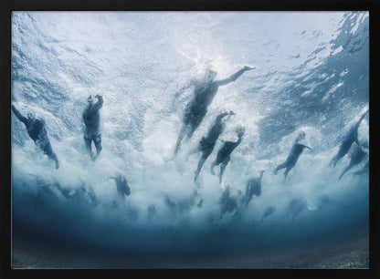 An underwater photograph looking up at a group of swimmers competing in a race. Their bodies are silhouetted against the bright surface of the turbulent blue water, captured mid-motion amidst a flurry of bubbles and foam. Print