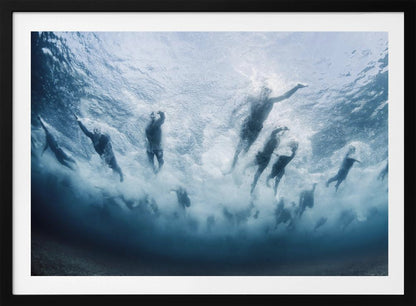An underwater photograph looking up at a group of swimmers competing in a race. Their bodies are silhouetted against the bright surface of the turbulent blue water, captured mid-motion amidst a flurry of bubbles and foam. Print