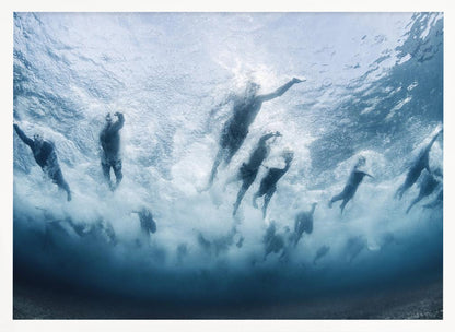 An underwater photograph looking up at a group of swimmers competing in a race. Their bodies are silhouetted against the bright surface of the turbulent blue water, captured mid-motion amidst a flurry of bubbles and foam. Print