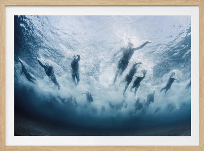 An underwater photograph looking up at a group of swimmers competing in a race. Their bodies are silhouetted against the bright surface of the turbulent blue water, captured mid-motion amidst a flurry of bubbles and foam. Print