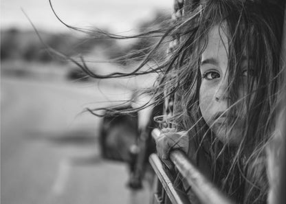 A close-up, black and white photograph of a young girl looking out of a moving vehicle. Her long hair is blowing wildly in the wind, partially obscuring her face as she peers intently at the camera with one eye. Her hand grips the edge of the open window. The background is blurred with motion, suggesting a journey. The photo is displayed in a silver frame. Poster