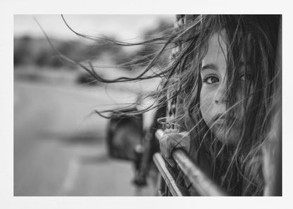 A close-up, black and white photograph of a young girl looking out of a moving vehicle. Her long hair is blowing wildly in the wind, partially obscuring her face as she peers intently at the camera with one eye. Her hand grips the edge of the open window. The background is blurred with motion, suggesting a journey. The photo is displayed in a silver frame. Poster