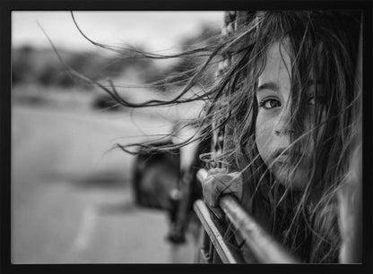 A close-up, black and white photograph of a young girl looking out of a moving vehicle. Her long hair is blowing wildly in the wind, partially obscuring her face as she peers intently at the camera with one eye. Her hand grips the edge of the open window. The background is blurred with motion, suggesting a journey. The photo is displayed in a silver frame. Poster
