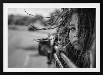A close-up, black and white photograph of a young girl looking out of a moving vehicle. Her long hair is blowing wildly in the wind, partially obscuring her face as she peers intently at the camera with one eye. Her hand grips the edge of the open window. The background is blurred with motion, suggesting a journey. The photo is displayed in a silver frame. Poster