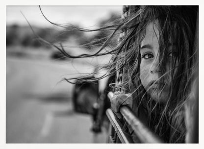 A close-up, black and white photograph of a young girl looking out of a moving vehicle. Her long hair is blowing wildly in the wind, partially obscuring her face as she peers intently at the camera with one eye. Her hand grips the edge of the open window. The background is blurred with motion, suggesting a journey. The photo is displayed in a silver frame. Poster
