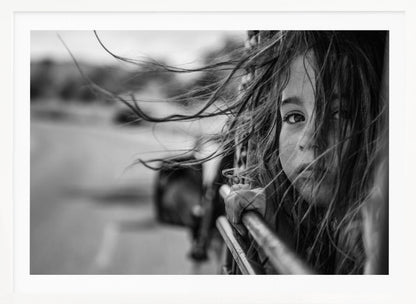 A close-up, black and white photograph of a young girl looking out of a moving vehicle. Her long hair is blowing wildly in the wind, partially obscuring her face as she peers intently at the camera with one eye. Her hand grips the edge of the open window. The background is blurred with motion, suggesting a journey. The photo is displayed in a silver frame. Poster