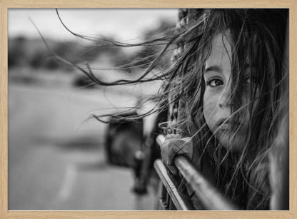 A close-up, black and white photograph of a young girl looking out of a moving vehicle. Her long hair is blowing wildly in the wind, partially obscuring her face as she peers intently at the camera with one eye. Her hand grips the edge of the open window. The background is blurred with motion, suggesting a journey. The photo is displayed in a silver frame. Poster
