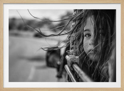 A close-up, black and white photograph of a young girl looking out of a moving vehicle. Her long hair is blowing wildly in the wind, partially obscuring her face as she peers intently at the camera with one eye. Her hand grips the edge of the open window. The background is blurred with motion, suggesting a journey. The photo is displayed in a silver frame. Poster
