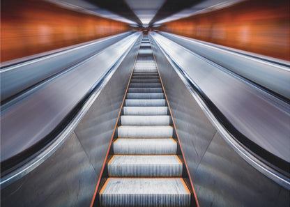 A symmetrical, low-angle photograph looking up a very long escalator, creating a strong sense of perspective towards a vanishing point. The orange walls are motion-blurred, suggesting speed, while the metallic steps in the center remain clear. Decor