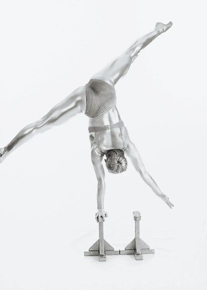 A black and white studio photograph of a muscular gymnast coated in silver paint, performing a dynamic one-armed handstand on a small block. Their legs are split wide apart in a display of strength and flexibility against a clean white background, all enclosed in a black frame. Decor