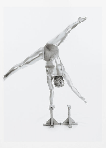 A black and white studio photograph of a muscular gymnast coated in silver paint, performing a dynamic one-armed handstand on a small block. Their legs are split wide apart in a display of strength and flexibility against a clean white background, all enclosed in a black frame. Decor