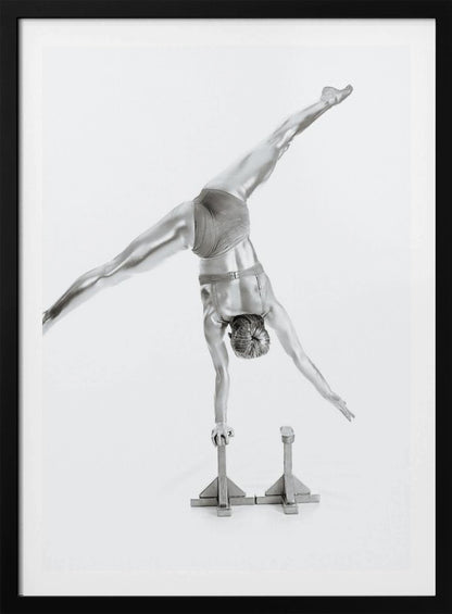 A black and white studio photograph of a muscular gymnast coated in silver paint, performing a dynamic one-armed handstand on a small block. Their legs are split wide apart in a display of strength and flexibility against a clean white background, all enclosed in a black frame. Decor