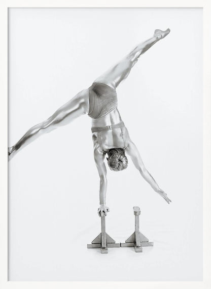 A black and white studio photograph of a muscular gymnast coated in silver paint, performing a dynamic one-armed handstand on a small block. Their legs are split wide apart in a display of strength and flexibility against a clean white background, all enclosed in a black frame. Decor