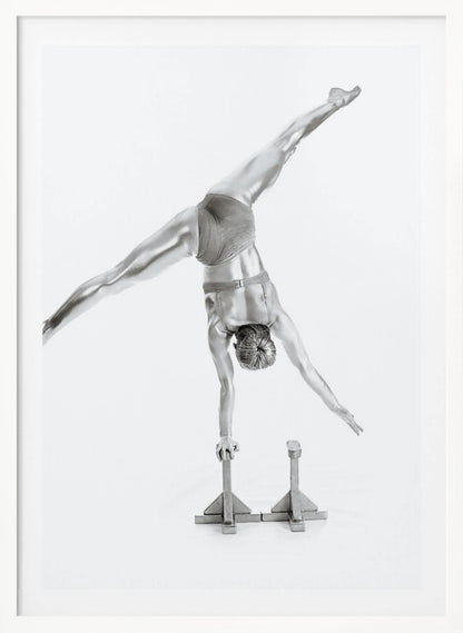 A black and white studio photograph of a muscular gymnast coated in silver paint, performing a dynamic one-armed handstand on a small block. Their legs are split wide apart in a display of strength and flexibility against a clean white background, all enclosed in a black frame. Decor