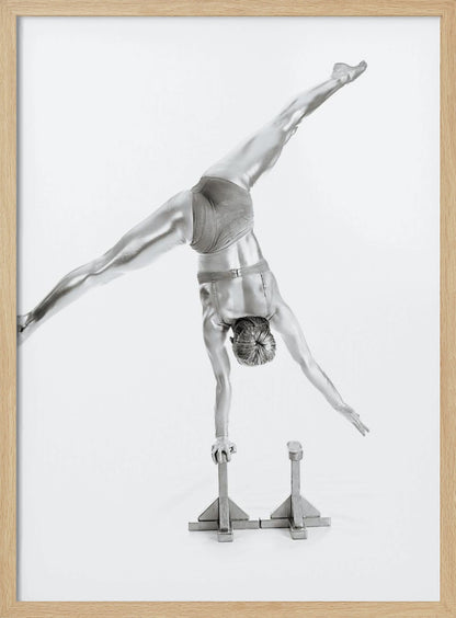 A black and white studio photograph of a muscular gymnast coated in silver paint, performing a dynamic one-armed handstand on a small block. Their legs are split wide apart in a display of strength and flexibility against a clean white background, all enclosed in a black frame. Decor