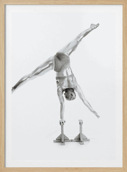 A black and white studio photograph of a muscular gymnast coated in silver paint, performing a dynamic one-armed handstand on a small block. Their legs are split wide apart in a display of strength and flexibility against a clean white background, all enclosed in a black frame. Decor