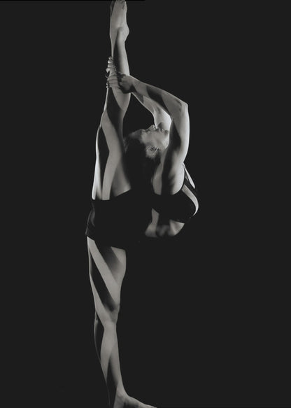 A dramatic black and white studio photograph of a female dancer performing a standing split. She balances on one leg while holding the other leg straight up vertically next to her head, arching her back and looking upwards. The strong side lighting creates a chiaroscuro effect, highlighting her musculature and form against a solid black background. Print