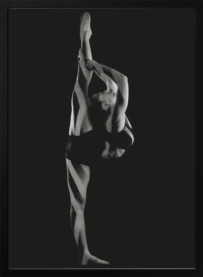 A dramatic black and white studio photograph of a female dancer performing a standing split. She balances on one leg while holding the other leg straight up vertically next to her head, arching her back and looking upwards. The strong side lighting creates a chiaroscuro effect, highlighting her musculature and form against a solid black background. Print
