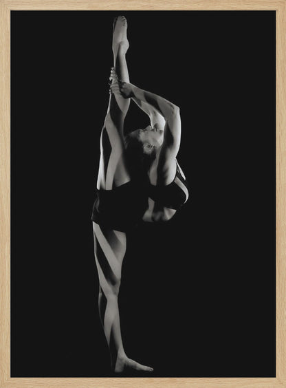 A dramatic black and white studio photograph of a female dancer performing a standing split. She balances on one leg while holding the other leg straight up vertically next to her head, arching her back and looking upwards. The strong side lighting creates a chiaroscuro effect, highlighting her musculature and form against a solid black background. Print