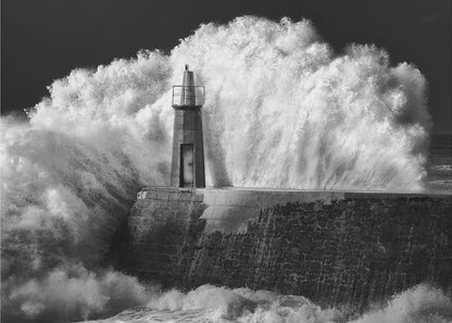 A dramatic black and white photograph of a massive wave crashing against a stone pier and a small lighthouse. The white foam of the wave towers over the structure, creating a powerful and dynamic scene against a dark sky, all enclosed in a silver frame. Poster