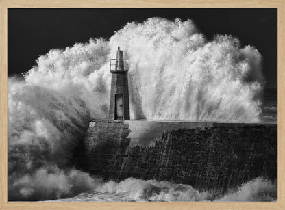 A dramatic black and white photograph of a massive wave crashing against a stone pier and a small lighthouse. The white foam of the wave towers over the structure, creating a powerful and dynamic scene against a dark sky, all enclosed in a silver frame. Poster