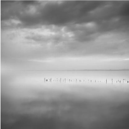 A minimalist black and white photograph in a light wood frame, depicting a calm, serene lake under a heavy, cloudy sky. The still water reflects the sky, with a row of weathered wooden posts breaking the surface along the horizon. Print