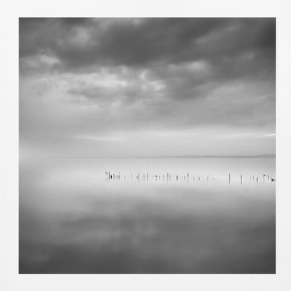A minimalist black and white photograph in a light wood frame, depicting a calm, serene lake under a heavy, cloudy sky. The still water reflects the sky, with a row of weathered wooden posts breaking the surface along the horizon. Print