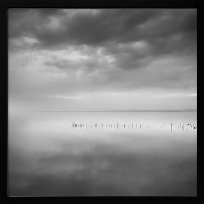 A minimalist black and white photograph in a light wood frame, depicting a calm, serene lake under a heavy, cloudy sky. The still water reflects the sky, with a row of weathered wooden posts breaking the surface along the horizon. Print