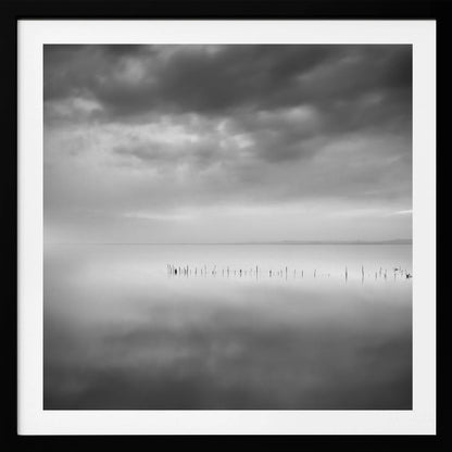 A minimalist black and white photograph in a light wood frame, depicting a calm, serene lake under a heavy, cloudy sky. The still water reflects the sky, with a row of weathered wooden posts breaking the surface along the horizon. Print