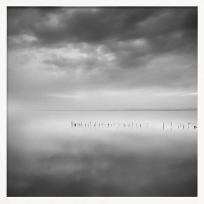A minimalist black and white photograph in a light wood frame, depicting a calm, serene lake under a heavy, cloudy sky. The still water reflects the sky, with a row of weathered wooden posts breaking the surface along the horizon. Print