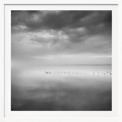 A minimalist black and white photograph in a light wood frame, depicting a calm, serene lake under a heavy, cloudy sky. The still water reflects the sky, with a row of weathered wooden posts breaking the surface along the horizon. Print