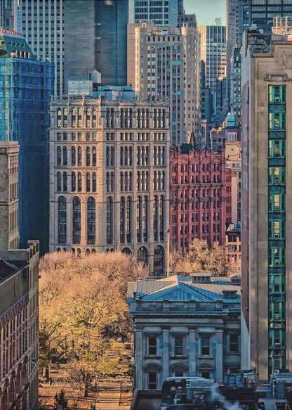 A high-angle view of a dense city skyline featuring a mix of old and new architectural styles. Golden light illuminates the autumn foliage of a park and the facades of several buildings, creating a warm contrast with the cool blue tones of the skyscrapers in the background. Decor