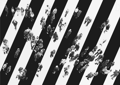 A striking black and white aerial photograph of dozens of people walking across a large, diagonally striped crosswalk. The high-contrast, graphic pattern of black and white stripes dominates the scene, with the small figures of pedestrians scattered across it, creating a sense of movement and urban anonymity. The image is presented within a simple silver frame. Print