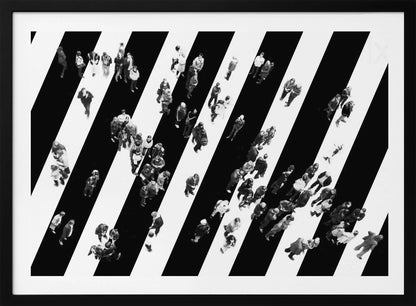 A striking black and white aerial photograph of dozens of people walking across a large, diagonally striped crosswalk. The high-contrast, graphic pattern of black and white stripes dominates the scene, with the small figures of pedestrians scattered across it, creating a sense of movement and urban anonymity. The image is presented within a simple silver frame. Print