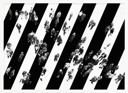 A striking black and white aerial photograph of dozens of people walking across a large, diagonally striped crosswalk. The high-contrast, graphic pattern of black and white stripes dominates the scene, with the small figures of pedestrians scattered across it, creating a sense of movement and urban anonymity. The image is presented within a simple silver frame. Print