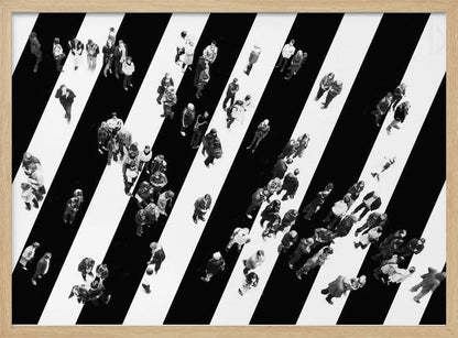A striking black and white aerial photograph of dozens of people walking across a large, diagonally striped crosswalk. The high-contrast, graphic pattern of black and white stripes dominates the scene, with the small figures of pedestrians scattered across it, creating a sense of movement and urban anonymity. The image is presented within a simple silver frame. Print