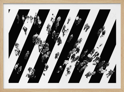 A striking black and white aerial photograph of dozens of people walking across a large, diagonally striped crosswalk. The high-contrast, graphic pattern of black and white stripes dominates the scene, with the small figures of pedestrians scattered across it, creating a sense of movement and urban anonymity. The image is presented within a simple silver frame. Print