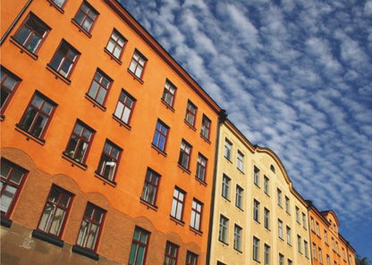 A low-angle shot of colorful buildings, predominantly a vibrant orange one, against a bright blue sky filled with a pattern of white, fluffy clouds. The image is presented within a silver-colored frame. Print