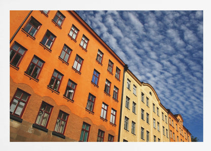 A low-angle shot of colorful buildings, predominantly a vibrant orange one, against a bright blue sky filled with a pattern of white, fluffy clouds. The image is presented within a silver-colored frame. Print