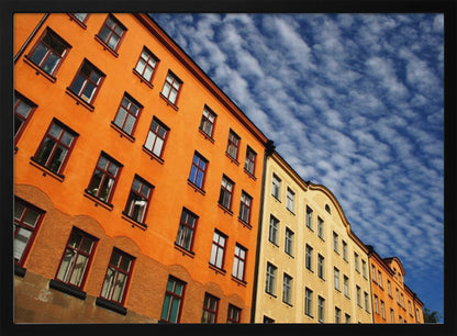 A low-angle shot of colorful buildings, predominantly a vibrant orange one, against a bright blue sky filled with a pattern of white, fluffy clouds. The image is presented within a silver-colored frame. Print