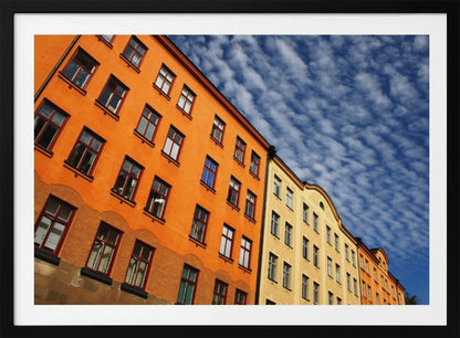 A low-angle shot of colorful buildings, predominantly a vibrant orange one, against a bright blue sky filled with a pattern of white, fluffy clouds. The image is presented within a silver-colored frame. Print