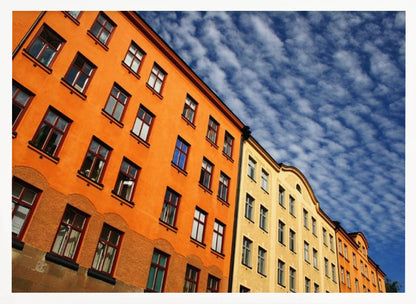 A low-angle shot of colorful buildings, predominantly a vibrant orange one, against a bright blue sky filled with a pattern of white, fluffy clouds. The image is presented within a silver-colored frame. Print