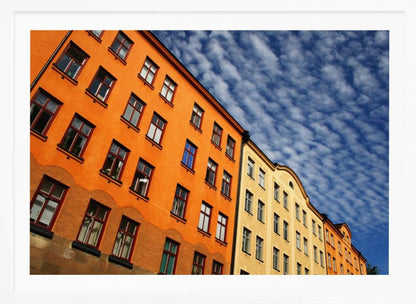 A low-angle shot of colorful buildings, predominantly a vibrant orange one, against a bright blue sky filled with a pattern of white, fluffy clouds. The image is presented within a silver-colored frame. Print