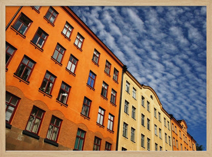 A low-angle shot of colorful buildings, predominantly a vibrant orange one, against a bright blue sky filled with a pattern of white, fluffy clouds. The image is presented within a silver-colored frame. Print