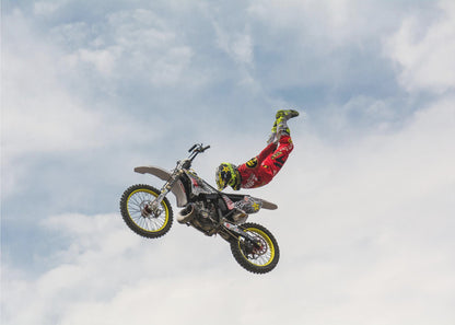 A motocross rider in red and yellow gear performs a high-flying stunt, arching their back while upside down in the air next to their dirt bike, set against a backdrop of a blue sky with white clouds and enclosed in a silver frame. Poster